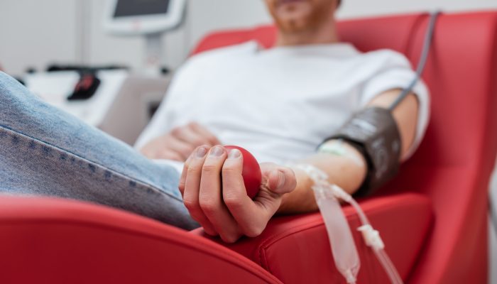 selective focus of rubber ball in hand of cropped volunteer with transfusion set sitting on comfortable medical chair near transfusion machine in blood donation center