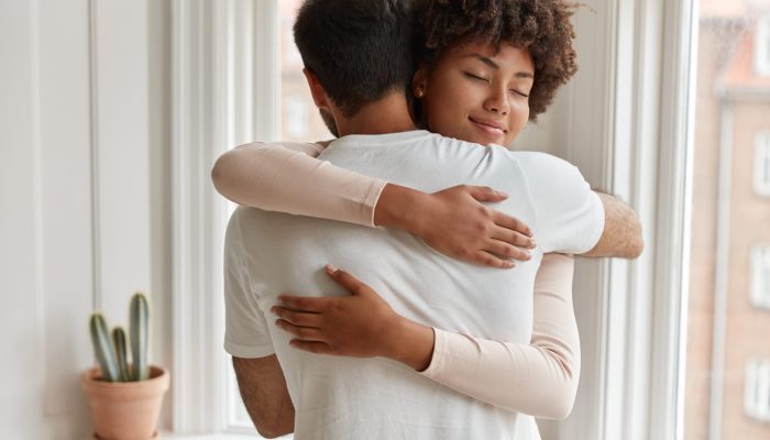 Pleased dark skinned young woman gives warm hug to her boyfriend, being pleased, pose near window, have romantic relationship, stand in cozy room. Husband and wife feel pleased and togetherness