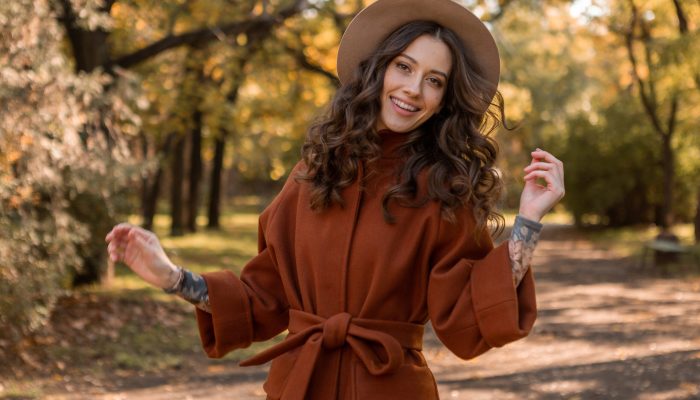 attractive stylish smiling skinny woman with curly hair walking in park dressed in warm brown coat, autumn trendy fashion street style, wearing hat