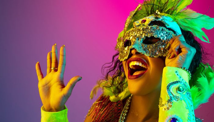 Close up. Beautiful young woman in carnival, stylish masquerade costume with feathers on gradient background in neon light. Concept of holidays celebration, festive time, dance, party, having fun.