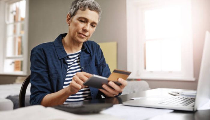 Shot of a senior woman using a credit and mobile while working on her finances at home