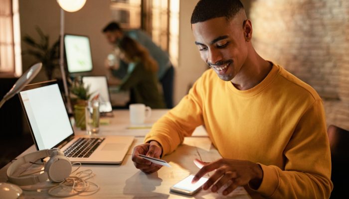 feliz-empresario-afro-americano-usando-telefone-celular-e-cartao-de-credito-de-pagamentos-online-enquanto-trabalhava-a-noite-no-escritorio