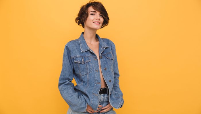 Portrait of a lovely young woman dressed in denim jacket posing while standing and looking at camera isolated over yellow background