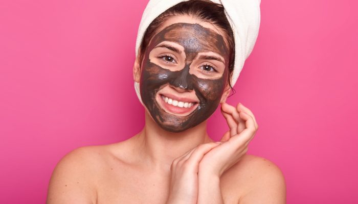 skin-care-concept-pleased-young-woman-with-toothy-smile-has-cocholate-facial-mask-isolated-pink-has-natural-beauty-wears-white-towel-has-nacked-shoulders-keeps-hands-near-face